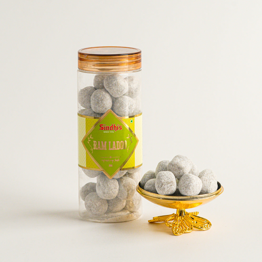 Jar of gray spherical candies with a gold lid and 'Sindhis' label, next to a small gold bowl filled with candies on a light background.