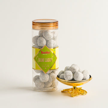 Jar of gray spherical candies with a gold lid and 'Sindhis' label, next to a small gold bowl filled with candies on a light background.