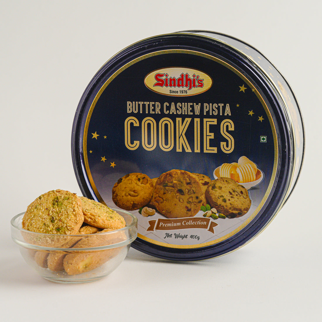 Cookies in a glass bowl with a container labeled 'Sindhi's Butter Cashew Pista Cookies' on a light gray background.