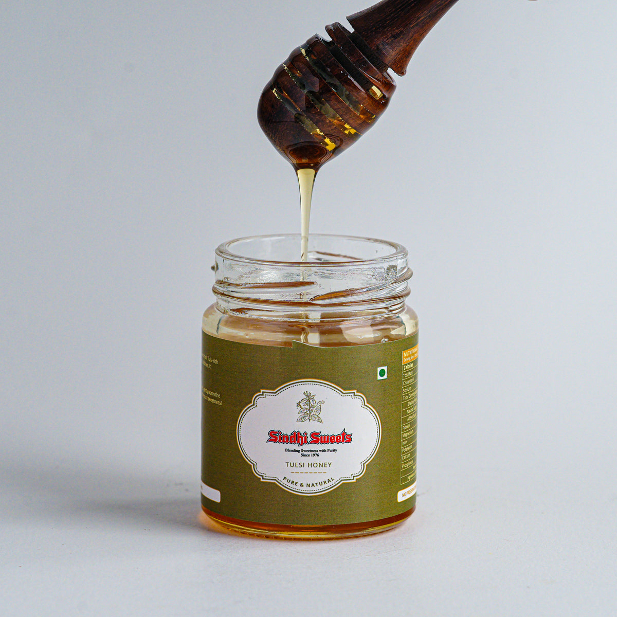 Honey being drizzled from a wooden dipper into a jar labeled 'Sindhi Sweets'.