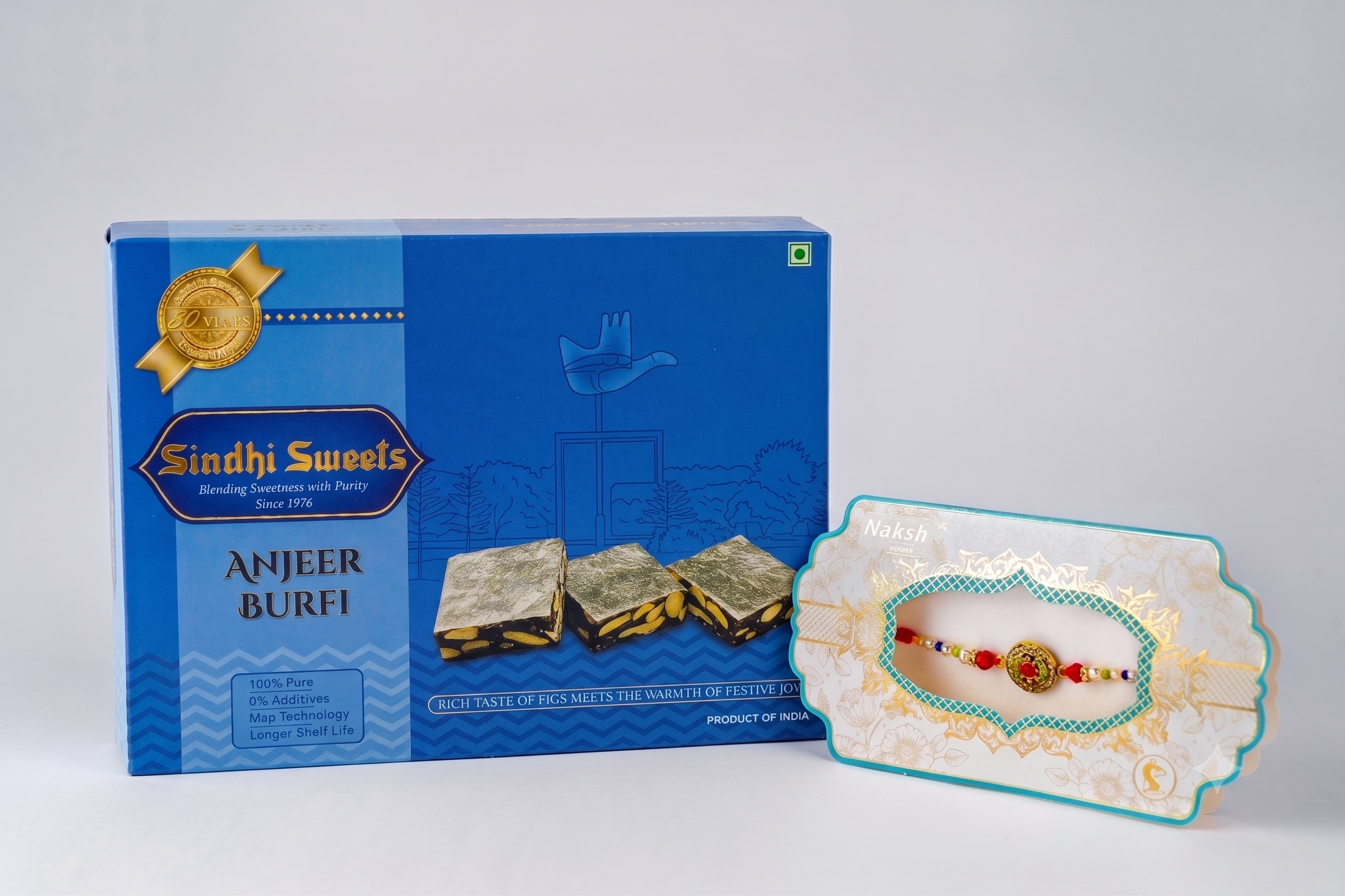 A blue box of Sindhi Sweets Anjeer Burfi next to a decorative white and teal plate holding a red and gold Rakhi, on a light gray background.