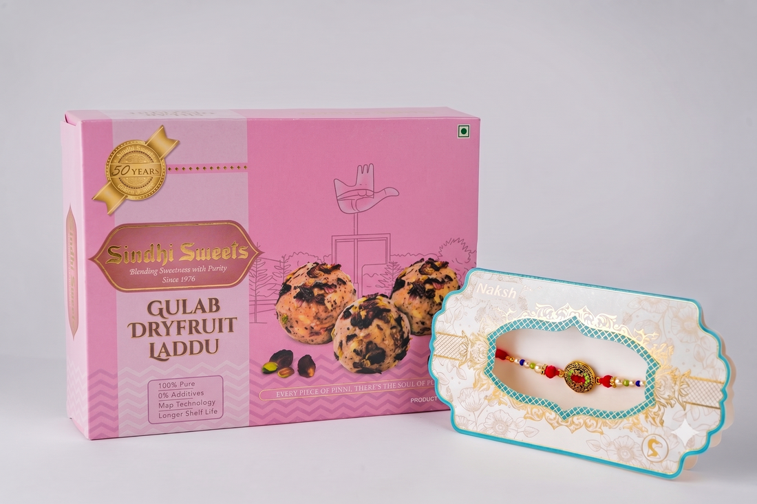 A pink box of Sindhi Sweets Gulab Dryfruit Laddu next to a decorative white and turquoise Rakhi with a gold ornament and red beads, set against a light gray background.