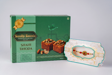 A green box of Sindhi Sweets Shahi Dhoda alongside a decorative white and teal gift tray holding a colorful beaded rakhi, photographed on a light gray studio background.