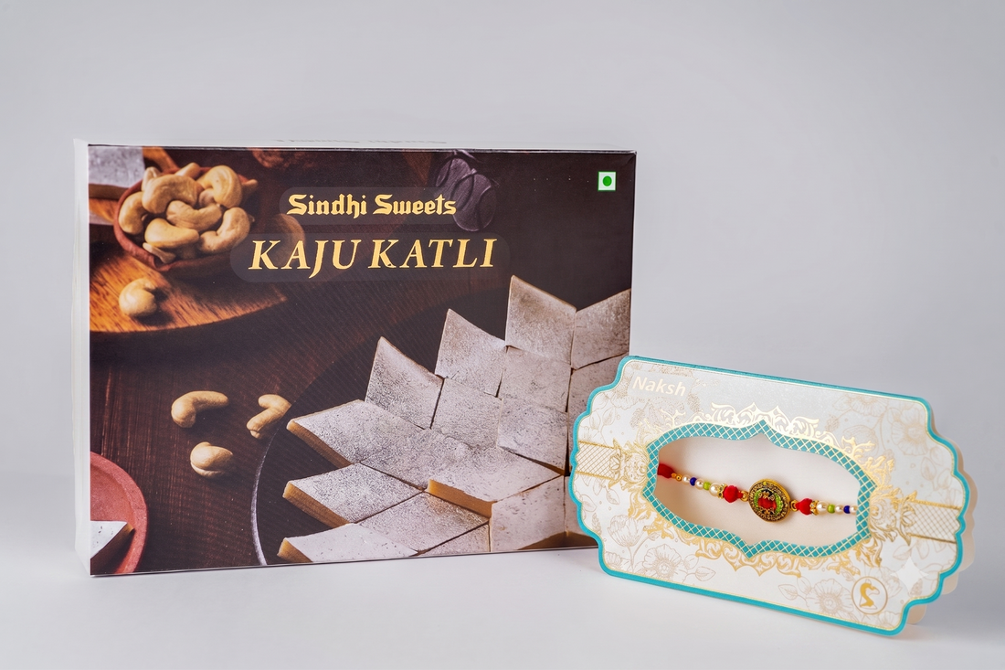 A box of Sindhi Sweets Kaju Katli next to a decorative gift card holding a small beaded Rakhi, photographed on a light gray studio background.