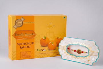 A yellow and orange box of Sindy Sweets Motichur Ladoo next to a decorated white and turquoise rakhi bracelet on a matching card, set against a plain light gray background.