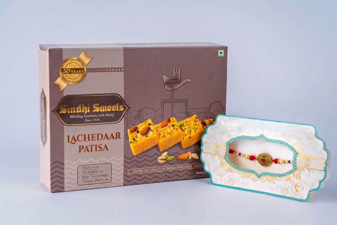 A box of Sandhi Sweets Lachedaar Patisa next to a decorative gift card holding a small Rakhi, photographed on a light gray studio background.