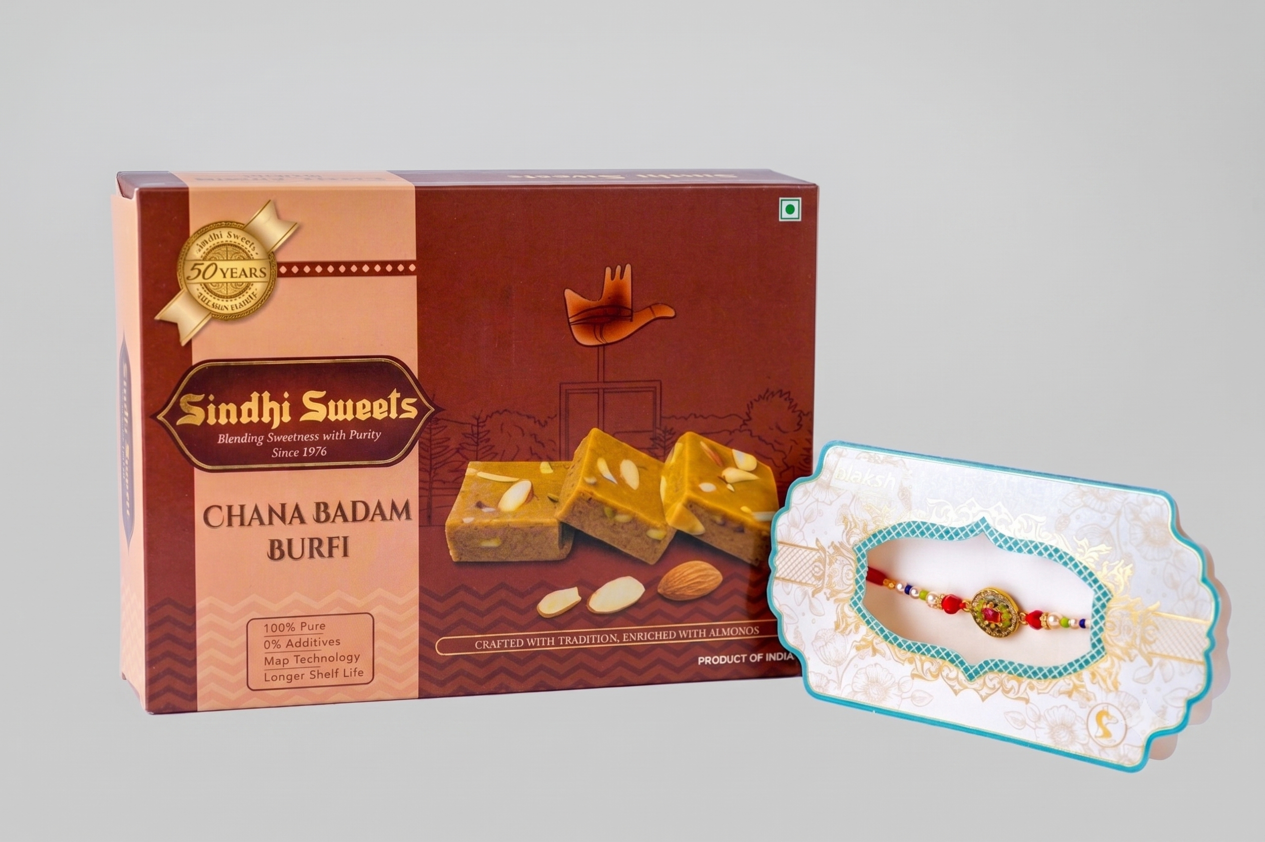 Sindhi Sweets Chana Badam Burfi box in dark brown and beige packaging, shown alongside a decorative white and turquoise Rakhi on a light gray background.